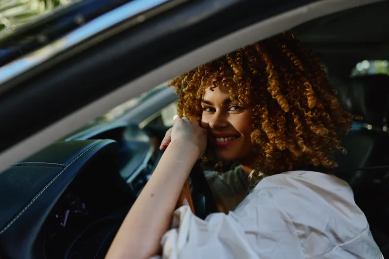 smiling woman with curly hair inside car steering wheel, natural light, casual clothing, warm and cheerful mood