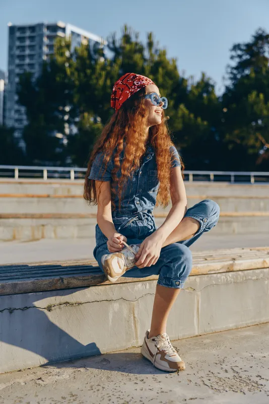Lifestyle woman in modern boho-western denim street style sitting outdoors, wearing red bandana and sunglasses, enjoying sunny day with film color tone, casual relaxed mood.