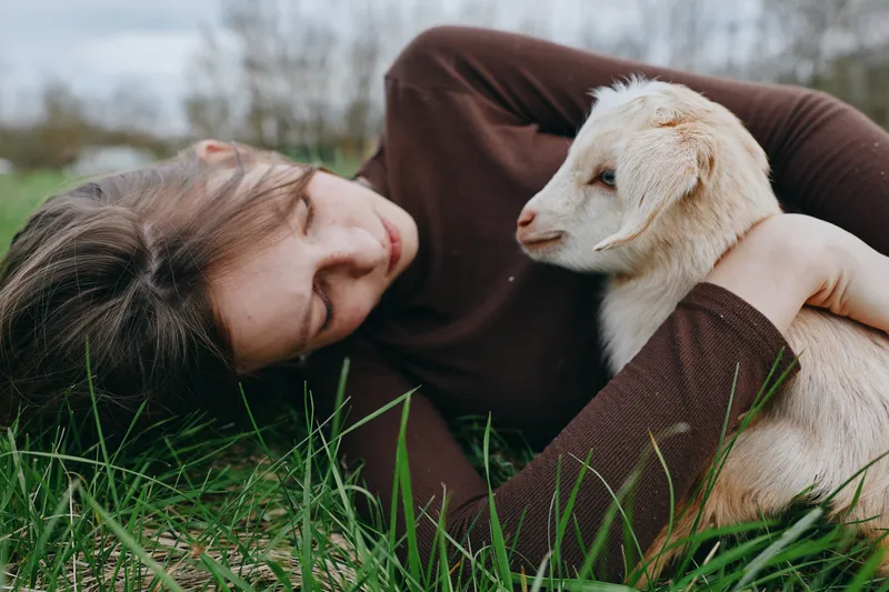 Relaxed woman lies on grass with a young goat beside her, sharing a quiet countryside moment, gentle bond, casual affection, peaceful mood, natural light, close connection, lifestyle scene outside,