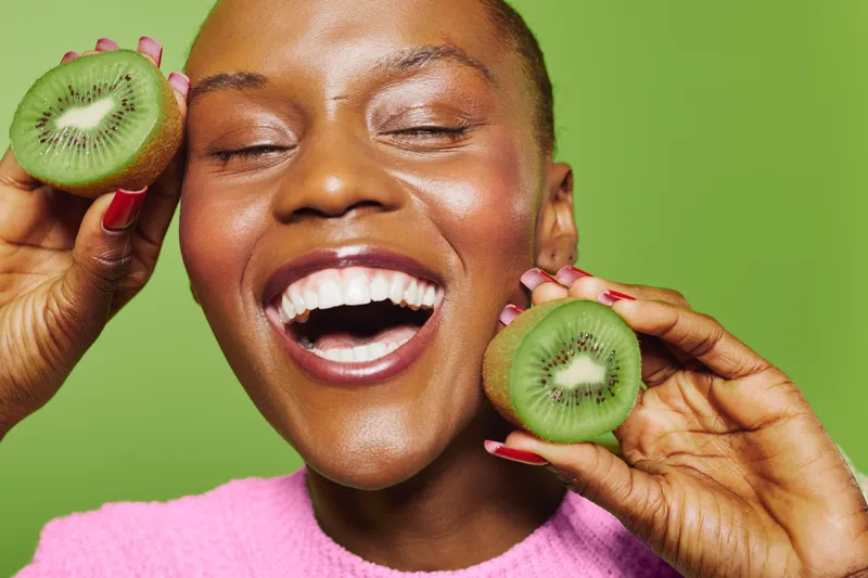 Happy African woman with dark skin laughs joyfully holding two kiwi halves near face. Vibrant green background and bright pink sweater create fresh and lively concept of healthy lifestyle and fruit.