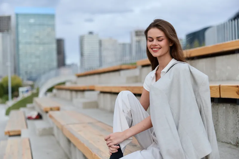 Business woman in a stylish outfit sitting on outdoor bleachers, smiling confidently with a modern cityscape in the background.