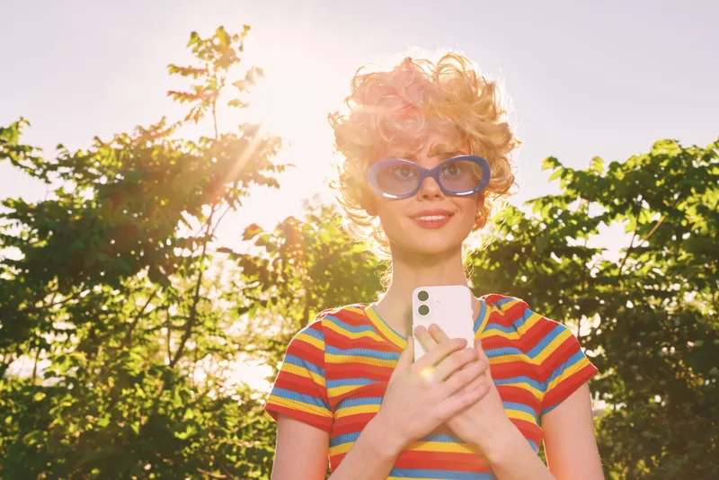 A stylish woman in a striped shirt and retro sunglasses outdoors, holding a smartphone. Bright sunlight and green trees behind her. Concept for lifestyle photography and nostalgia.