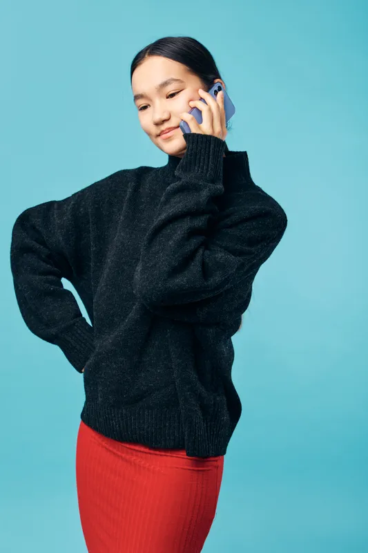 Young woman in black sweater and red skirt talking on mobile phone with hand on hip standing against blue background. Portrait of confident female using smartphone for communication.