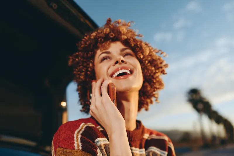 Woman lifestyle outdoors cheerful laughing, curly hair, phone near face, warm sunlit street light, relaxed confident expression, casual sweater, soft breeze, natural colors, shallow depth