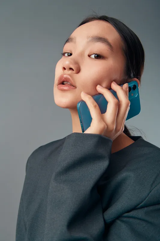 Young woman holding smartphone to her ear with contemplative expression wearing grey long sleeve top on neutral background. Closeup portrait, communication and modern technology concept.
