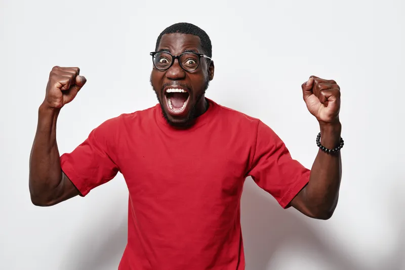 Excited black man with dark skin wearing glasses and red t shirt celebrating success with raised fists and open mouth on white background. Emotion, joy and victory concept.