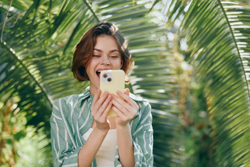 Young woman smiles while using a smartphone outdoors among lush tropical plants, a bright sunlit scene.
