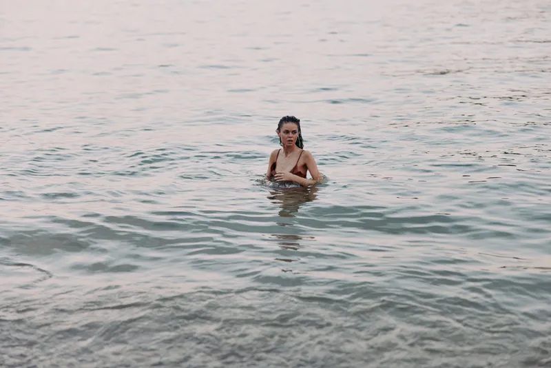 A lone woman stands waist-deep in clear water, gentle waves ripple around her as warm light glints on the surface, creating a serene beach scene.
