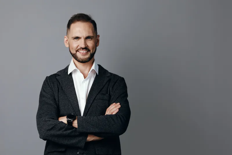 A confident business professional man stands with arms crossed, showing warm emotions against a solid colored background, conveying leadership, focus, and determination in a corporate setting.