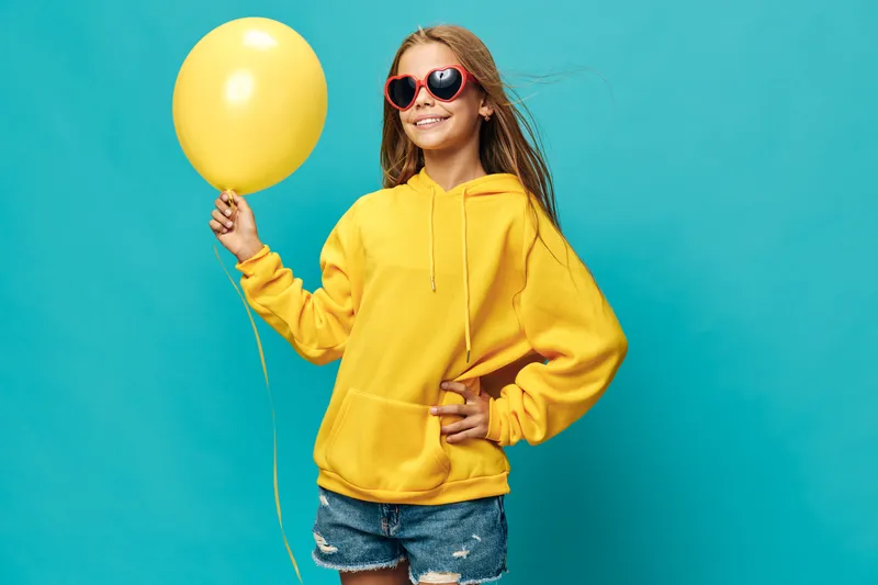 Girl in yellow hoodie and sunglasses stands with yellow balloon smiling against blue background. Child with casual outfit, summer style, and positive joy for celebration or fun time.