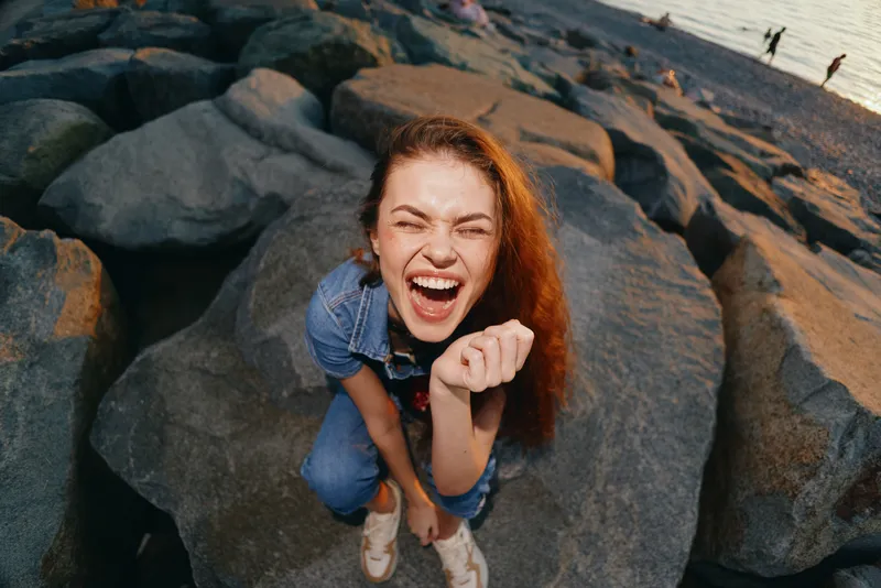 Lifestyle woman in modern boho-western denim street style laughing joyfully on rocky beach with warm film color tones and casual relaxed mood