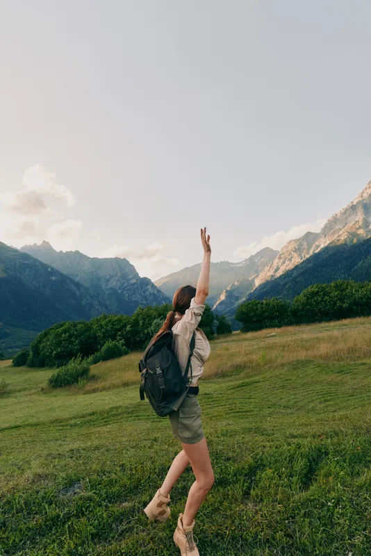 A young woman with a backpack raises her arm while walking through a green meadow surrounded by mountains under a clear sky, expressing freedom and adventure.