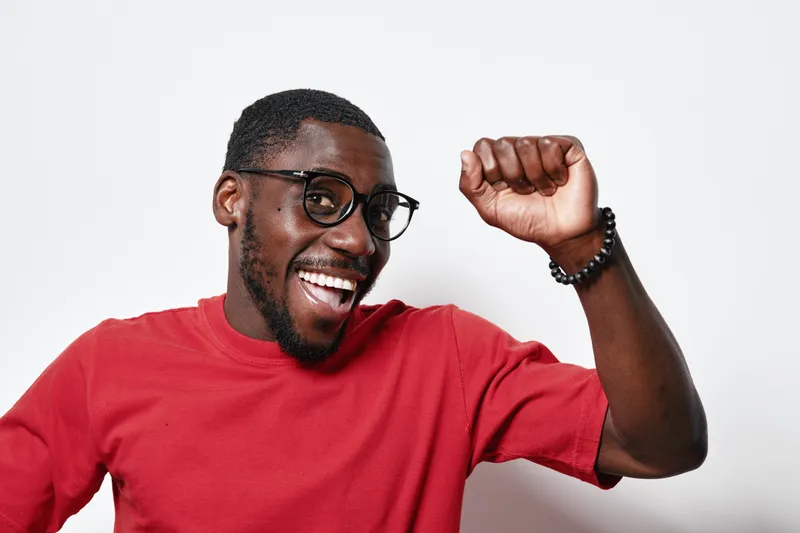 African American man with dark skin wearing glasses and red shirt smiling and raising fist in celebration on white background. Happy male portrait with positive emotion.