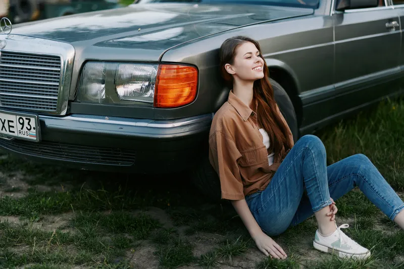 Young woman enjoying a sunny day sitting beside a vintage car, showcasing casual fashion and a relaxed expression in a natural outdoor setting.
