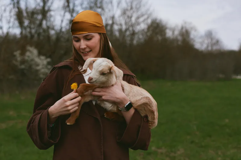 A woman in a brown coat smiles softly while cradling a young goat kid in a green field, offering a dandelion. A warm lifestyle moment highlighting animal bonding and rural gentleness.