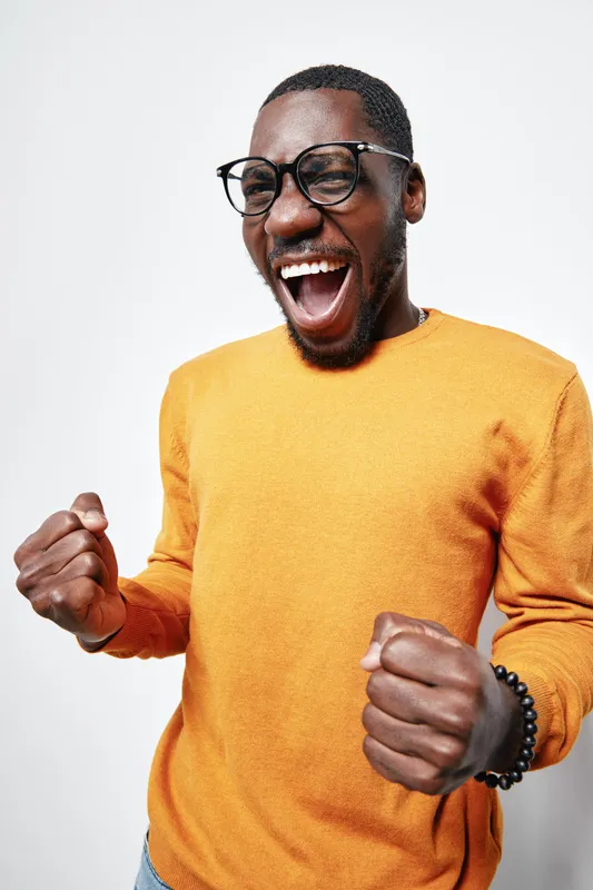 African American man with dark skin wearing glasses and orange sweater expressing excitement with clenched fists and open mouth on white background. Happy, energetic, casual style.
