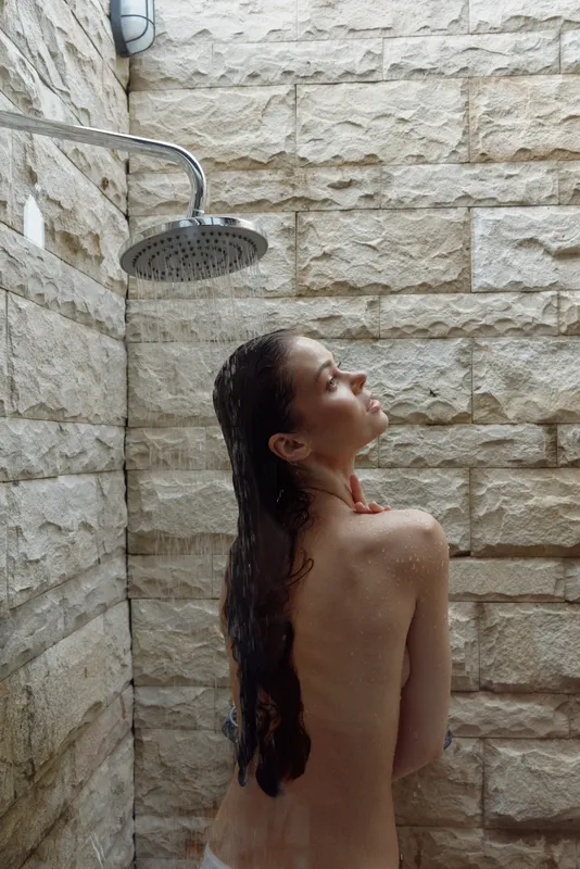 young woman enjoying outdoor shower with wet hair, natural stone wall background, calm and relaxing moment in soft daylight