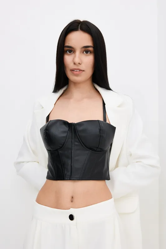 young woman wearing black leather bustier and white blazer, posing confidently against plain white background, modern fashion style