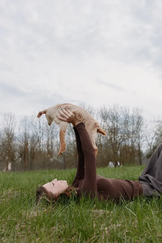 Young woman lying on grass lifts a baby goat above her with a joyful expression in a natural outdoor setting during a cloudy day