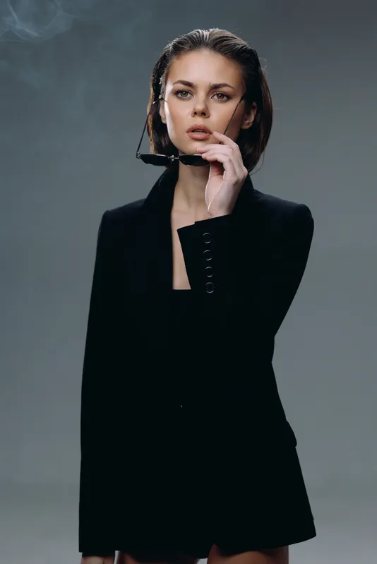 Stylish woman in black holding sunglasses with smoky background, confident and elegant expression