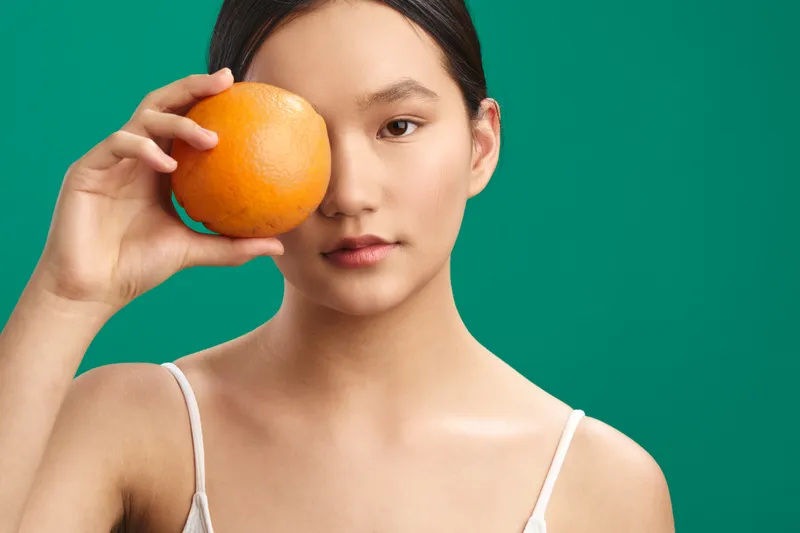 Asian woman holds an orange near her eye against a bright green backdrop, presenting a playful, bold portrait with soft lighting and minimal styling.