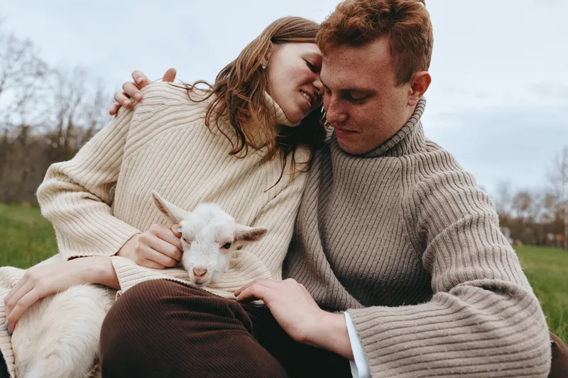 Smiling couple in knit sweaters cuddle a goat kid outdoors on a grassy field, warm mood, rustic setting, lifestyle moment, gentle affection, companionship, nature, happiness