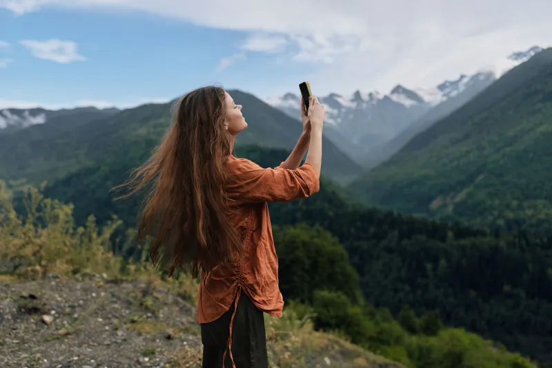 Woman takes a selfie in a rugged mountains landscape, outdoor scene conveying travel and nature vibes as wind moves her hair, capturing a vivid horizon with a smartphone