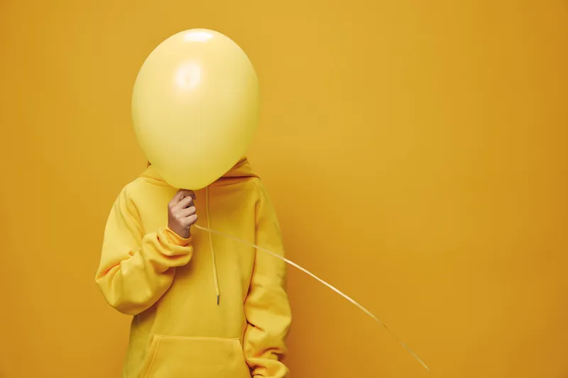 Person holding yellow balloon covering face wearing yellow hoodie on yellow background. Casual outfit with bright monochrome style for creative or playful concept in studio.