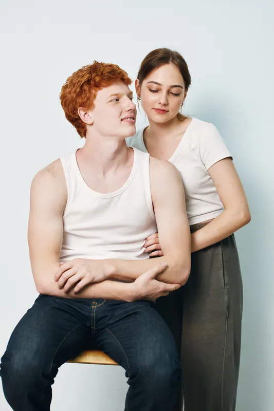 Young couple posing together in casual clothes against plain background. Man with red hair sitting and woman standing with gentle expression and arms around waist.