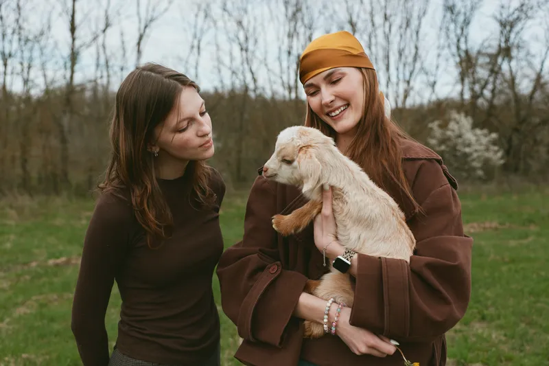 Two women outdoors share a gentle moment with a smiling goat kid, warm jackets, natural light, a relaxed lifestyle scene filled with friendship, nature, and rustic charm in a green field