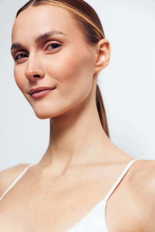 Close-up of a smiling woman with clear skin and tied hair wearing white top. Soft lighting highlights natural beauty and healthy complexion. Concept of skincare and wellness.