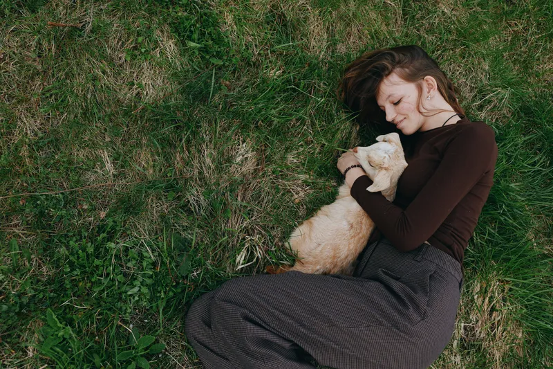 Woman lies on grass outdoors, holding a light cat with tenderness, enjoying a calm, warm moment in natural light, a simple lifestyle scene that emphasizes affection, relaxation, and peaceful