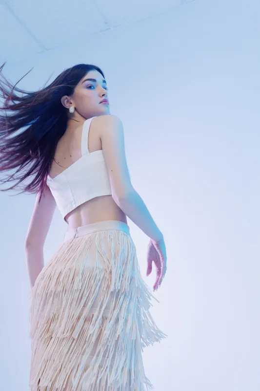 Young woman beauty fashion model poses with flowing hair wearing a white top and fringed skirt on a light blue background. Concept of style and elegance.