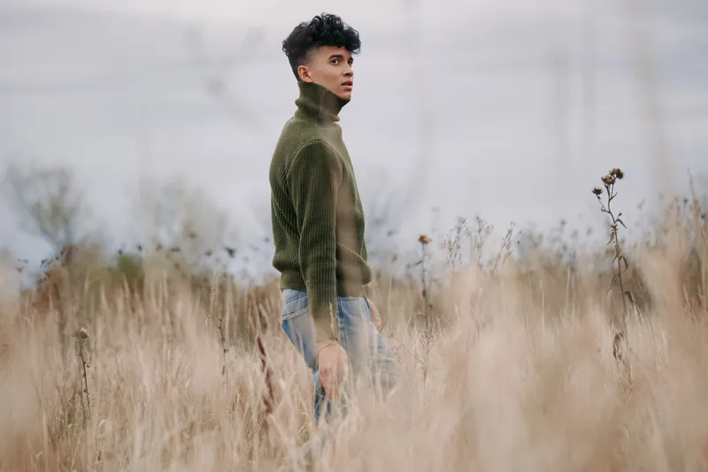 Candid portrait of a young man standing in tall dry grass, wearing a textured green sweater, conveying a genuine, natural presence and believable everyday style.