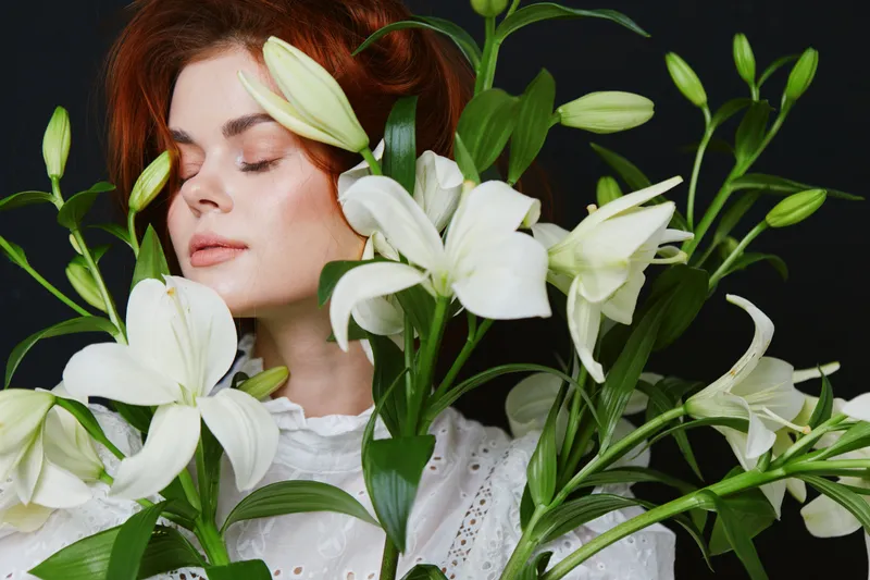 Woman with white lilies, soft expression, wearing a lace blouse against a dark background, symbolizing beauty and serenity in floral aesthetics