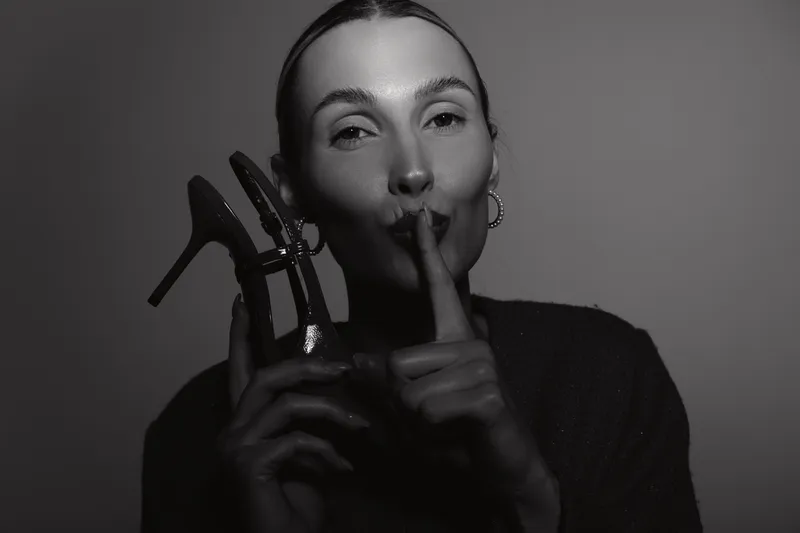Elegant caucasian woman with light skin holding high heel shoes and making silence gesture in black and white portrait. Concept of secrecy and fashion.