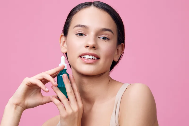 Young woman holding a skincare serum bottle, showcasing a healthy skincare routine, against a soft pink background