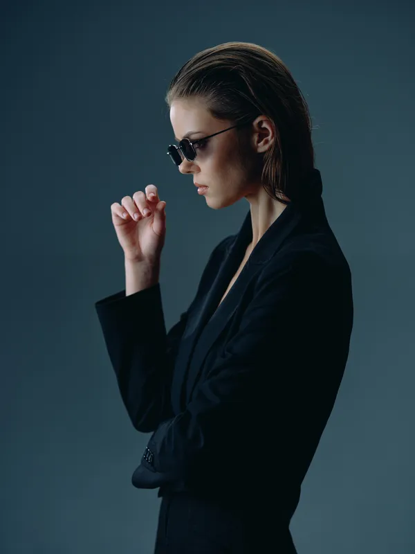 thoughtful woman in sunglasses wearing black suit posing against dark background