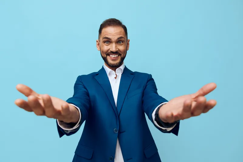 A confident business professional man stands against a solid blue background with open arms, conveying warmth, approachability and enthusiasm for any client oriented project.