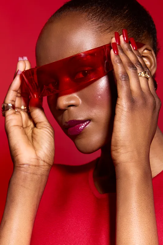Closeup portrait of elegant African woman with dark skin wearing red futuristic sunglasses and red outfit. Fashion concept with glossy makeup and stylish manicure on red background.