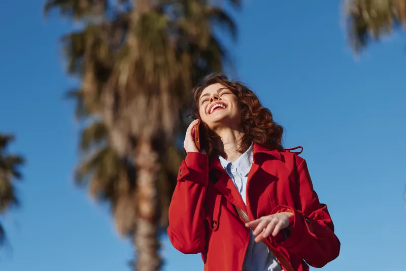 Young woman with curly hair smiles joyfully while talking on the phone outdoors under clear blue sky with blurred palm trees in the background.