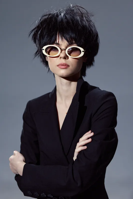 Stylish woman with short black hair and unique sunglasses wearing a black blazer with confident expression on gray background