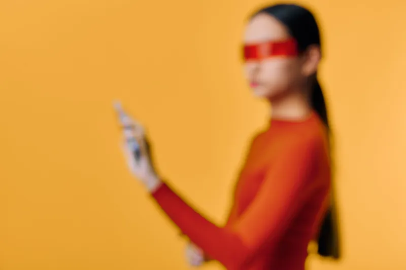 Blurred woman in red top and futuristic red glasses holding smartphone against bright orange background for technology and lifestyle concept in digital communication.