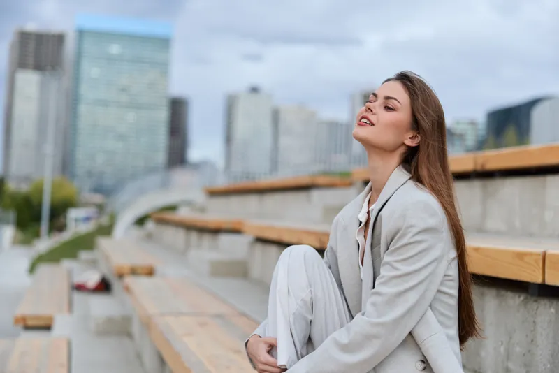 Young woman in light gray suit relaxing outdoors with urban skyline background, casual confident pose, autumn city lifestyle concept