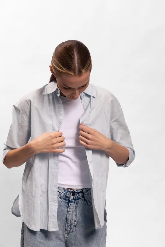 Young woman with brown hair wearing casual light shirt and white top looks down while buttoning shirt. Studio portrait on white background. Concept of fashion, casual style, and clothing.