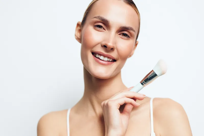 Smiling woman with natural makeup holding a makeup brush on white background. Concept of beauty, skincare and cosmetic application. Soft lighting highlights fresh and healthy skin tone.