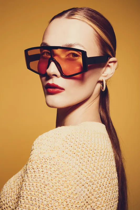 Stylish woman with long hair wearing oversized orange sunglasses and knitted sweater poses against yellow background. Fashion portrait with bold makeup and modern accessories.