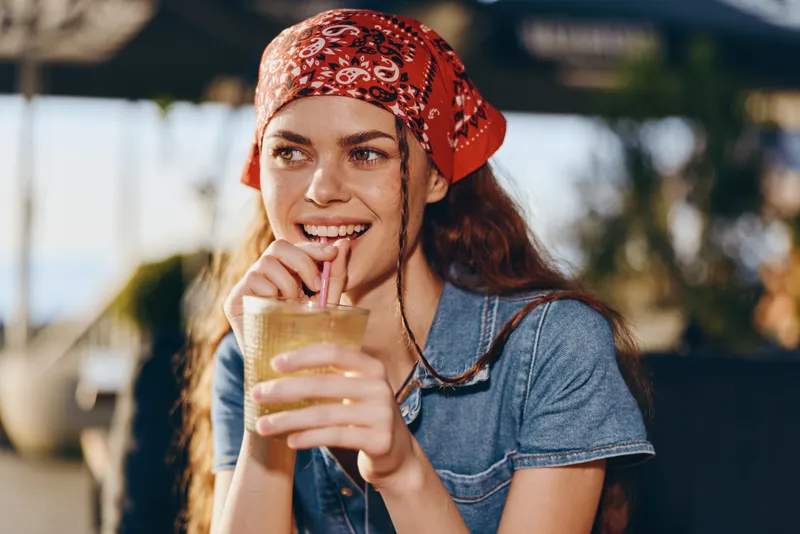 Smiling woman drinking cold beverage outdoors wearing red bandana and casual denim shirt, enjoying sunny day with relaxed and happy mood.
