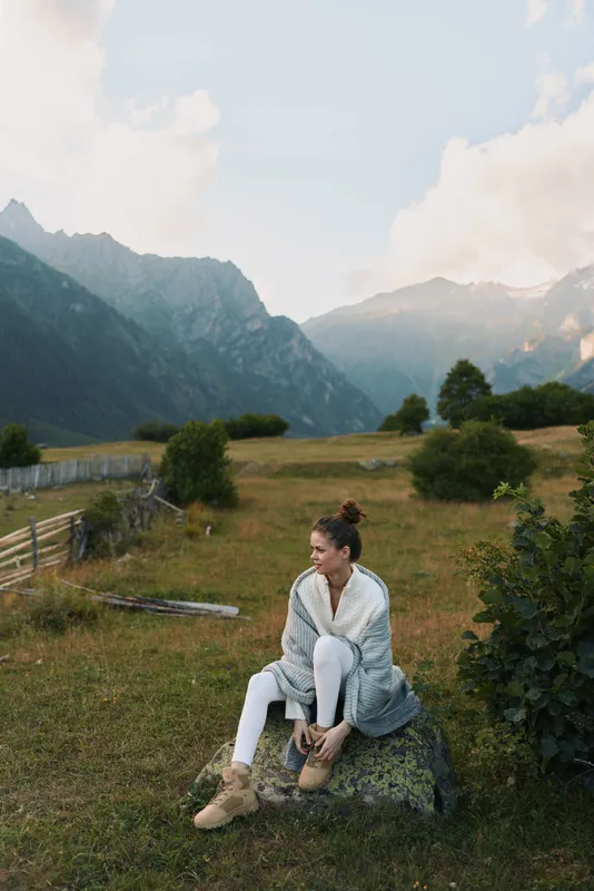 Young woman in a cozy sweater and boots sits on a rock in a mountain meadow enjoying the peaceful natural landscape during a cool day.