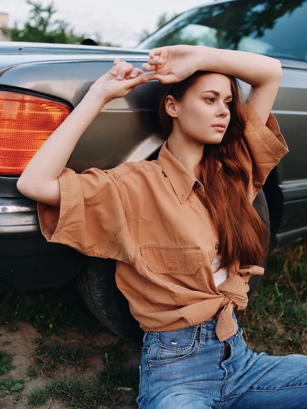 Young woman in a casual brown shirt and denim jeans leaning against a car, expressing a relaxed and thoughtful mood in a sunlit outdoor setting.